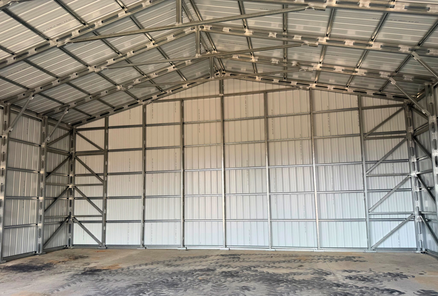 Inside back view of 40'x40'x15' steel shop features ladder style truss design, vertical roof, 14 Ga. frame, 30PSF ground snow load, fully enclosed with vertical siding, one 36" x 80" walk-in door, and two 12' x 14' overhead door frame outs on the end.